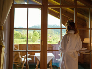 Side view of attractive female in robe is approaching the window at home and taking a sip of coffee. Woman in a winter hotel admiring the view savoring cup of coffee.