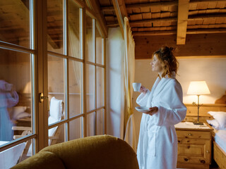 Obraz premium Tourism vacation and a woman standing in white robe at hotel window overlooking the view. Female enjoying the coffee by the window of chalet.