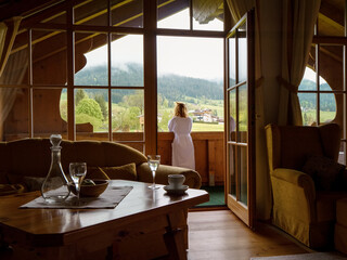 Tourism vacation back view of beautiful woman standing in white robe on hotel balcony overlooking the landscape. Large view of female admiring nature by panoramic window of hotel.