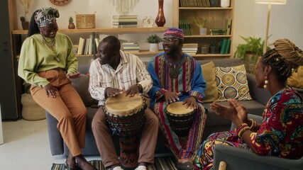 Four Black young friends dressed in vibrant clothing celebrating cultural holiday at home, two men playing traditional African drums while women clapping to beat and tapping rhythm - Powered by Adobe