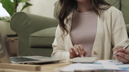 Selection focuses on the hand. Close up of young woman sitting and using a calculator for analyzing financial data together with a laptop computer about her family business in living room at her home. - Powered by Adobe