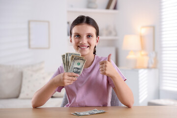 Smiling little girl with dollar banknotes showing thumbs up at wooden table indoors. Pocket money and responsibility