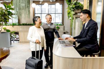 Happy senior Asian couple checking in at hotel on holiday vacation with young male receptionist....