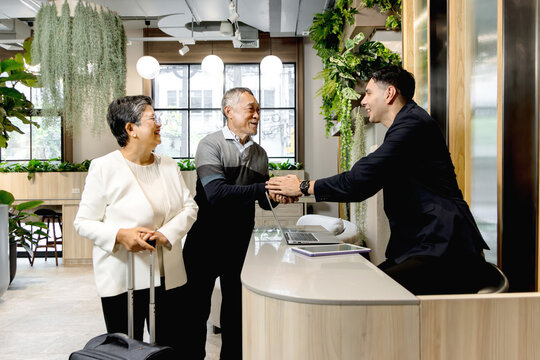 Happy senior Asian couple checking in at hotel on holiday vacation with young male receptionist. Receptionist man in suit shaking hands with Elderly traveler customer at hotel reception counter desk.