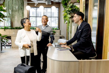 Happy senior Asian couple checking in at hotel on holiday vacation with young male receptionist....