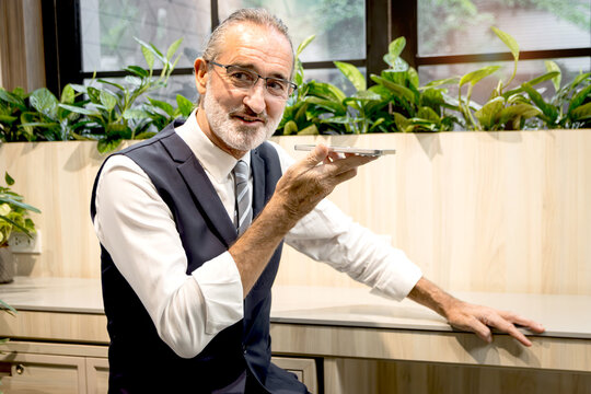 Happy senior businessman talks on smartphone at modern office. Elderly man connects with AI technology at co working space office workplace. Mature manager uses mobile phone to control remotely.