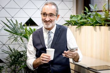 Happy senior businessman drinking coffee and holding smartphone at modern office. Elderly man sipping coffee at co working space office workplace during a break. Mature manager taking a break.