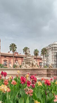 Hispalis Fountain in Seville, one of the city's most beautiful fountains, surrounded by colorful flowers and historical buildings. Timelapse hyperlapse with a cloudy sky in the background.
