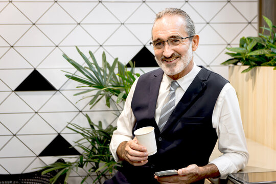 Happy senior businessman drinking coffee and holding smartphone at modern office. Elderly man sipping coffee at co working space office workplace during a break. Mature manager taking a break.