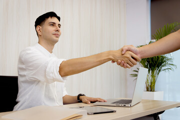 Happy smiling young businessman has handshake after finish business deal with partner. Successful business worker greeting with handshake with other while sitting and working on laptop computer.