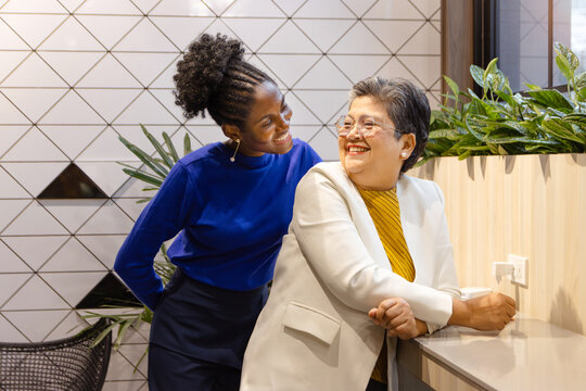Happy Asian senior businesswoman and African female officer with curly hair stand at office, look at each other. Beautiful business staff workers working together. Diversity and harmony in workplace