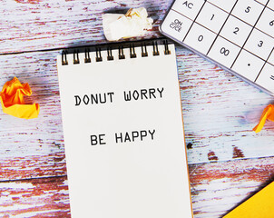 A notebook displays the phrase DONUT WORRY BE HAPPY. It rests near a calculator and crumpled papers on a weathered wooden table. A motivational message for a relaxed mindset