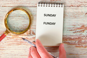 A hand holds a patterned pencil near a notepad that says SUNDAY FUNDAY. A magnifying glass sits nearby, all on a light, aged, wooden table surface