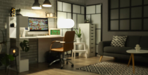 Blurred view of dark programmer's office with workplace, sofa and glowing lamps at night