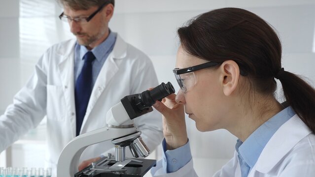 Female scientist wearing protective glasses looking through a microscope during medical research in laboratory with her male colleague working in the background. Medicine and science concept