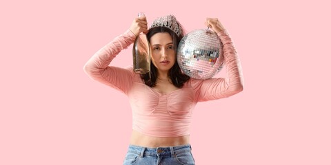 Young woman with hangover after party holding disco ball and bottle of champagne on pink background