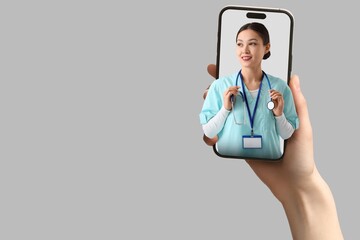 Patient holding smartphone with female Asian doctor looking out of screen against grey background. Concept of telemedicine