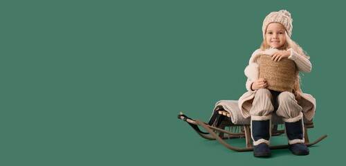 Cute little girl in winter clothes and with basket sitting on sled against green background with...