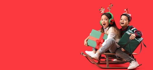 Emotional little children with Christmas gifts and sled on red background with space for text