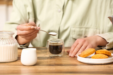Woman adding spoon of sugar into cup of coffee at table in kitchen. Closeup