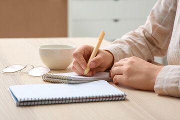 Woman with eyeglasses and cup of coffee writing something in notebook at workplace, closeup