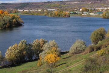 Peaceful autumn landscape with a calm river, colorful trees, and a grazing cow on a green hillside under a cloudy sky