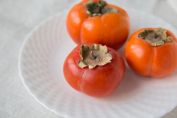 Three persimmons on a white plate on a light-colored textile background