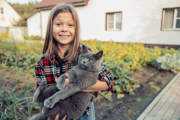 A young Caucasian girl with long brown hair smiles while holding a gray cat in her arms. They are in a garden near a house, surrounded by plants and greenery.