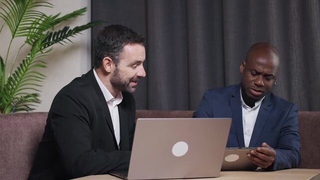 Two professional men engaged in a collaborative discussion while working on a laptop at a modern office space, showcasing teamwork and innovation in a business environment	