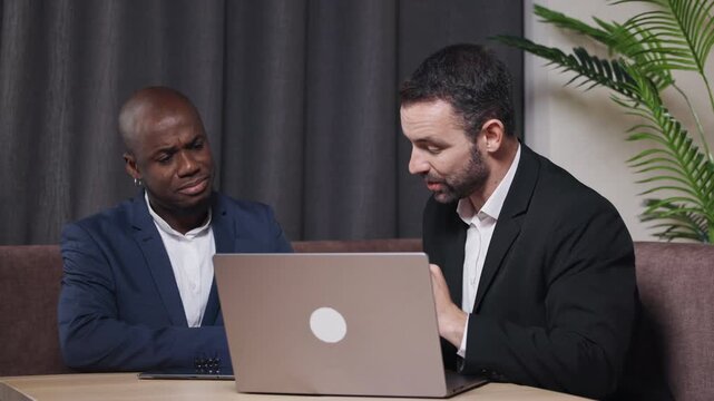 Two professional men engaged in a collaborative discussion while working on a laptop at a modern office space, showcasing teamwork and innovation in a business environment	