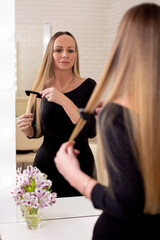 
Young beautiful woman brushing her long healthy blonde hair and looking at the mirror in hairdressing salon
