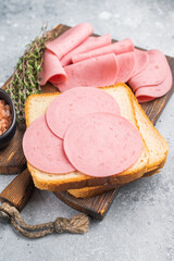 Slices of pink bologna lunch meat on toasted white bread with rock salt and thyme on a wooden board. Grey background with copy space. © Mironov