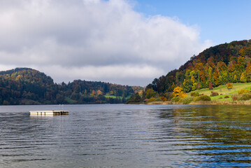 Autumn lake landscape. Fall season forest and mountains on a sunny day. Scenic Swiss view of a calm lake with a wooden raft and vibrant autumn trees under a cloudy sky. Turlersee, Switzerland.