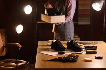 Boots with shoe polishes and brushes on table against male shoemaker in dark workshop