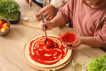 Little girl with her father applying tomato sauce on pizza dough at table in kitchen, closeup