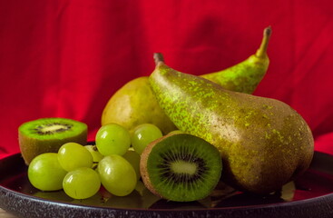 fresh green fruits on red background: ripe pear, green grapes, and a sliced kiwi on a plate