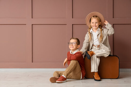 Stylish little children with suitcase sitting near brown wall - Powered by Adobe