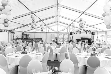 Eine helle, große Festzelt-Location ist für eine Feier festlich eingedeckt, wobei runde Tische...