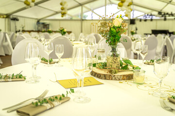 Eine helle, große Festzelt-Location ist für eine Feier festlich eingedeckt, wobei runde Tische mit strahlend weißen Tischdecken und Überwürfen über Stühlen dominieren. Die Dekoration ist in weiß-golde