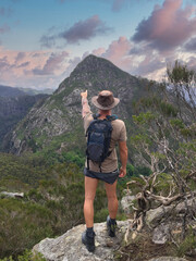Obraz premium Back view of a lone Brazilian hiker with a hat and backpack standing on a rocky outcrop, pointing toward a distant mountain peak in the forested highlands of Australia under a colorful sky.