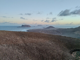 Aerial view of a volcanic cone rising from the arid landscape of La Graciosa Island, Canary Islands, with the Atlantic Ocean in the background.

