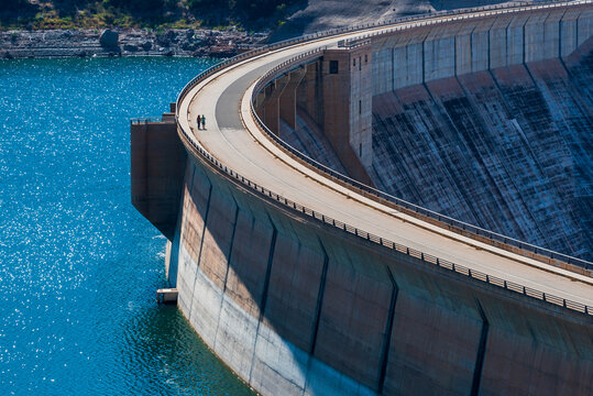 Massive curved concrete dam with small couple walking on the closed road, aerial side view