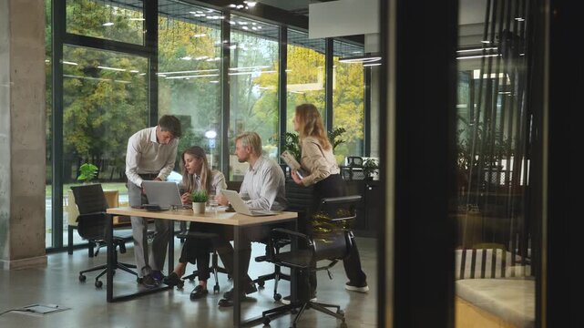 A view through a glass partition shows a modern, busy office interior. Business people are working at desk