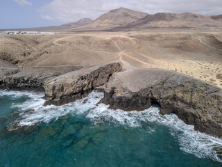 Aerial view of rugged cliffs and golden sand beach near Playa Blanca, Lanzarote, with turquoise Atlantic waters and arid volcanic terrain.
