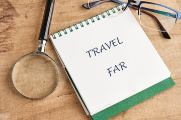 A notebook with TRAVEL FAR written on it sits beside spectacles and a magnifier on a wooden surface, suggesting preparation for a vacation