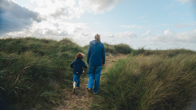 A mother and her child walk hand in hand through grassy sand dunes under a partly cloudy sky. The scene captures a peaceful moment of connection and exploration in nature, with soft natural light and 