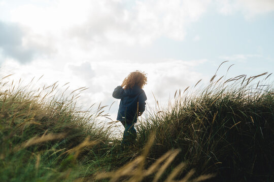 A young child with curly hair walks through tall dune grass under a bright, cloudy sky. Captured from a low angle, the photo conveys a sense of wonder and adventure in nature. The wind gently moves th - Powered by Adobe