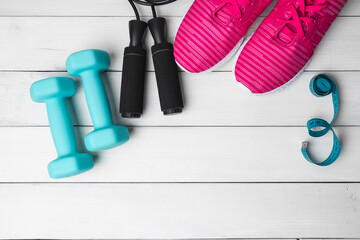 Top view of pink sneakers, blue dumbbells, jump rope and measuring tape on white wooden background with copy space, fitness equipment flat lay concept
