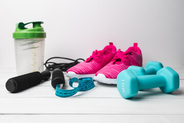 Pink sneakers, blue dumbbells, jump rope, shaker and measuring tape on white wooden background with copy space, fitness equipment and active lifestyle concept
