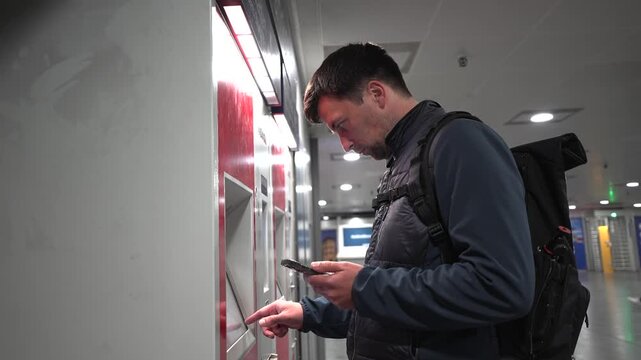 A man with a backpack uses a red Deutsche Bahn ticket machine in Munich, Germany. He holds a smartphone and selects a public transport ticket on the touchscreen at the train station.